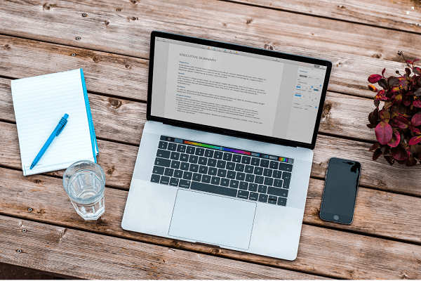 Laptop on a wooden table with phone, notebook, pen, water glass, and plant
